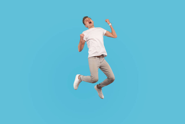 Full length, excited successful man in white t-shirt and casual pants jumping with happiness and shouting yes, I did it, feeling inspired and energetic. indoor studio shot isolated on blue background - Valokuva, kuva