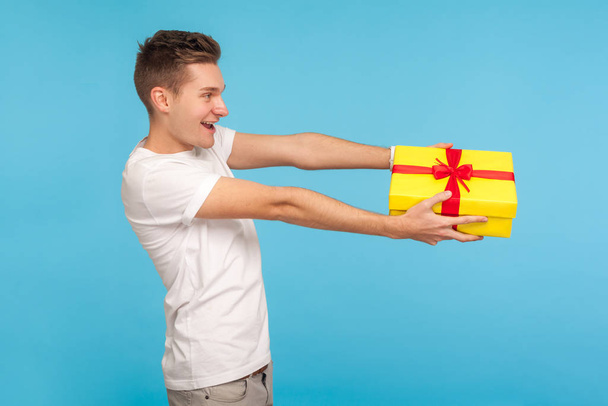 Charity and generosity. Side view of happy man in white t-shirt giving gift box and smiling with good-natured expression, offering birthday present. indoor studio shot isolated on blue background - Foto, Imagem