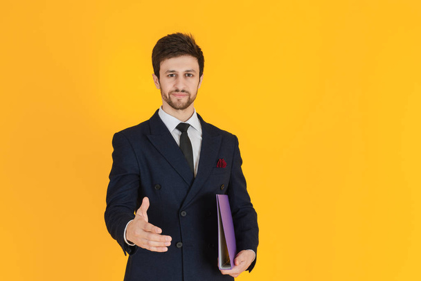 Een jonge zakenman in een pak houdt een documentenbestand vast en reikt een hand uit om de hand te houden op de gele geïsoleerde achtergrond. Zakelijk imago voor banners. - Foto, afbeelding