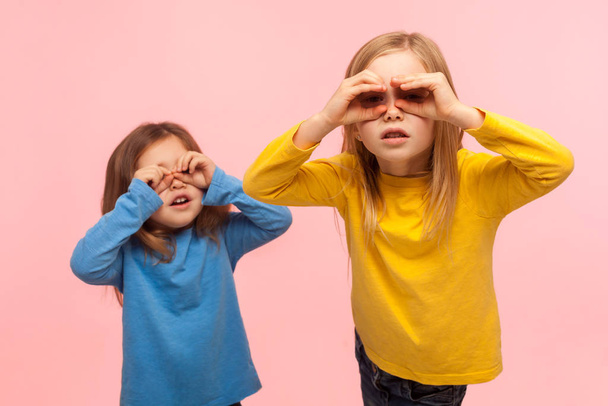 Adorable, curious, nosy two little girls looking through fingers imitating binoculars, exploring world, discovering something new with interested gaze. indoor studio shot isolated on pink background - Photo, Image