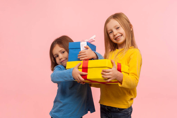 Holiday present for kids. Two happy little girls holding gifts together and smiling at camera, children embracing christmas presents, birthday surprise. indoor studio shot isolated on pink background - Foto, Bild