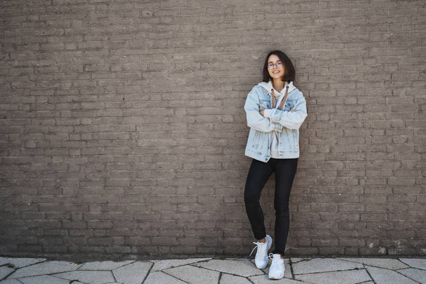 Education, lifestyle and modern generation concept. Confident happy young woman in denim jacket, glasses, lean brick wall and smiling, cross hands over chest confident, sending CV found job - Foto, Imagem