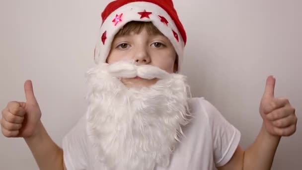 girl with artificial beard in Santa Claus hat raises thumbs up on white backdrop - Footage, Video