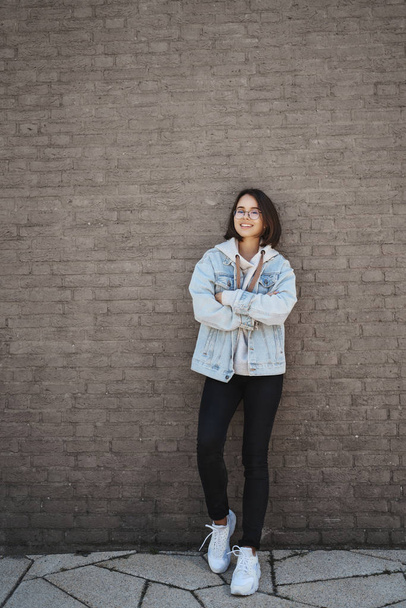Modern lifestyle and people concept. Portrait of happy young hipster girl in denim jacket, glasses, lean casually brick wall and cross hands chest, express confidence and chill, smiling camera - Foto, Imagem