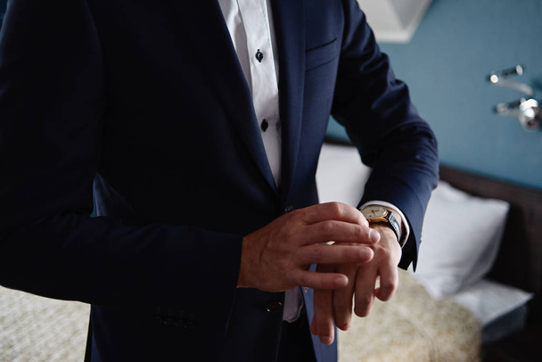 Geschäftsmann, der drinnen auf seine Armbanduhr schaut, kopiert den Raum. Mann im blauen Anzug, der die Zeit von Luxus-Armbanduhren kontrolliert. Beobachten Sie auf der Hand. Hochzeitsvorbereitungen für den Bräutigam - Foto, Bild