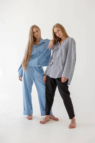 two pretty young twin sisters with long blond hair posing on white background in oversize clothes. Fashion photoshoot - Fotó, kép