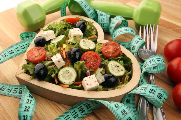 Authentischer frischer Salat in einer hölzernen herzförmigen Tasse mit Hanteln, Maßband auf dem Tisch. gesunde Lebensweise, gute Gesundheit Konzept mit frischen Zutaten - Foto, Bild
