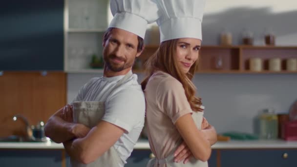 Portrait of chef and assistant in hats standing back to back on kitchen - Záběry, video