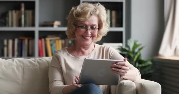 Happy mature woman using digital tablet computer sitting on couch - Felvétel, videó