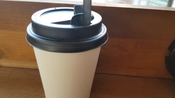 There is a white paper Cup of coffee on a wooden table in the cafe, and through the glass you can see the street with a blurred focus - Video, Çekim