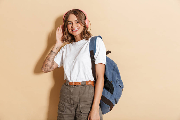 Portrait of a smiling schoolgirl carrying backpack standing isolated over beige background, listening to music with wireless headphones - 写真・画像