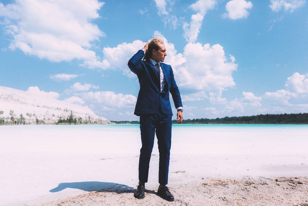 Blond krullende man loopt op het strand van kristalheldere zee. Gekleed zakenpak, blauwe kleur. Zomer, trendy. Reisthema - Foto, afbeelding