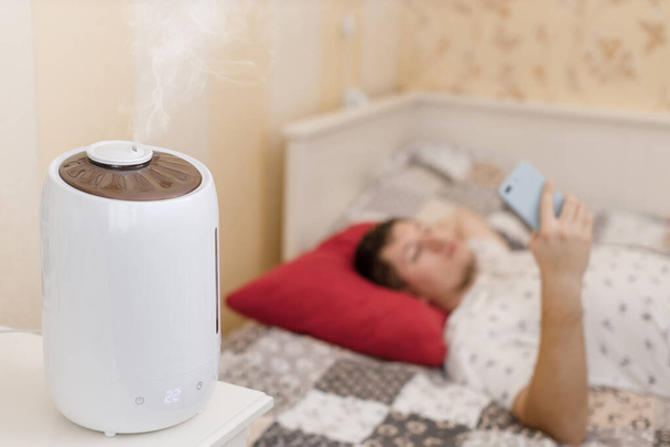 White humidifier spreading steam. Humidification of the dry air in sleeping room. Selective focus on vapor. On the background young man surfing in web. Air purity and healthcare concept. Home interior - Photo, Image