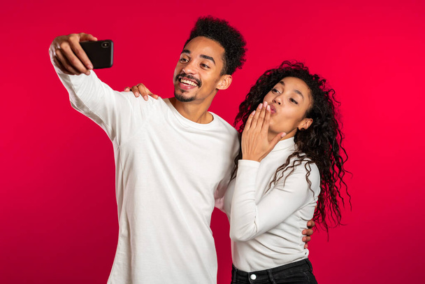 Young african american couple in white making selfie with smartphone on red studio background.Lovers match, love, holidays, happiness concept - Foto, imagen