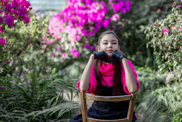 lovely girl sitting on a chair against a background of brightly flowering plants - Photo, Image