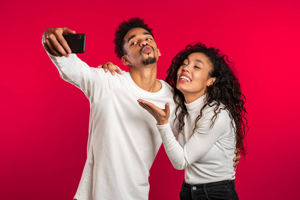 Jeune couple afro-américain en blanc faisant selfie avec smartphone sur fond de studio rouge.Lovers match, amour, vacances, concept de bonheur
 - Photo, image