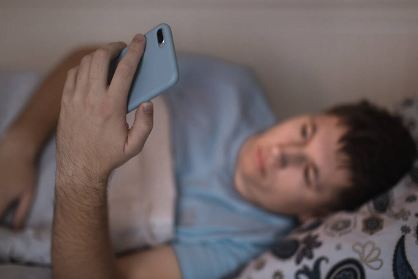 Sleepy tired male awaked late at night in bed and surfing in web. Social media addiction, dependency on a cell phone, negative effect on the eyes, sleeplessness concept. Selective focus on smartphone. - Foto, Imagen