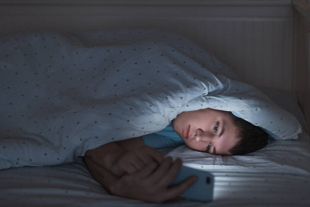 Slaperig depressief tiener surfen op het internet op zijn mobiele telefoon liggend op bed in het donker. Web verslaving, smartphone afhankelijkheid, internet val. Slapeloosheid, nomofobie, slaapstoornis concept. - Foto, afbeelding