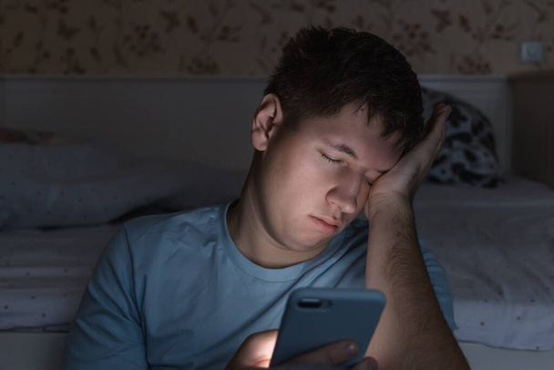 Adult sleepy man awake late at night in bed surfing in web, can not fall asleep/ sleepy tired, social media addiction, dependency on cell phone, half-closed tired eyes, insomnia, sleeplessness concept - Fotoğraf, Görsel