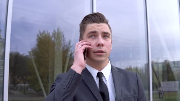 A young businessman in a suit speaks on the phone. Serious man stands in front of a mirror business center - Video