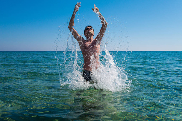 Junger Mann mit Sonnenbrille springt im Sommer aus dem Meer und verursacht Spritzer, Rhodos, Griechenland. Lizenzfreies Archivfoto. - Foto, Bild