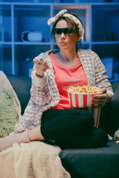 Mujer en un sofá blando comiendo palomitas de maíz, mirando la pantalla del televisor en gafas 3D. Mujer comiendo palomitas de maíz en vasos 3d
. - Foto, imagen