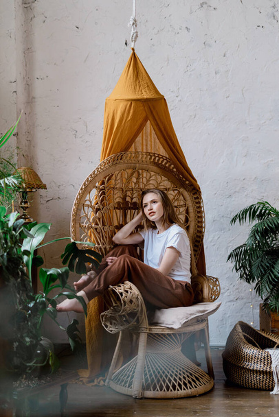 Photo verticale d'une jeune femme adulte rêveuse et souriante assise sur une chaise en osier dans une pièce confortable, passant la journée à la maison, avec des plantes vertes dans des pots de fleurs
 - Photo, image