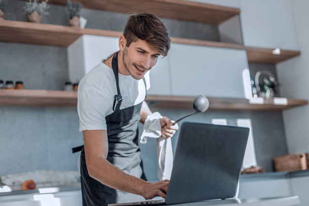 young man looking at recipe in laptop while cooking dinner - Фото, изображение