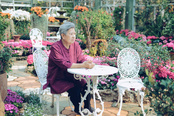 old elder woman resting in flower garden. asian elderly female relaxing in park. senior leisure lifestyle - Zdjęcie, obraz