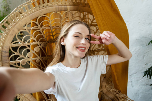Веселая молодая женщина проводит свободное время дома, моргает глазом, широко улыбается и делает селфи фото, показывая знак победы в камере
 - Фото, изображение