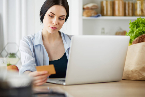 Hymyilevä nainen verkkokaupoissa tietokoneella ja luottokortilla keittiössä - Valokuva, kuva