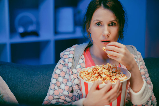 Portret van de vrouw met popcorn die op de bank zit en iets engs kijkt terwijl ze popcorn eet en bang is - Foto, afbeelding