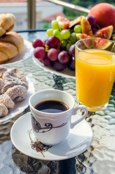 Healthy breakfast on the table with a cup of coffee, a glass of fresh orange juice, cookies in the foreground. Croissants and fruits: grapes, figs, peach in the background. Lunch on terrace outdoor. - Foto, Bild