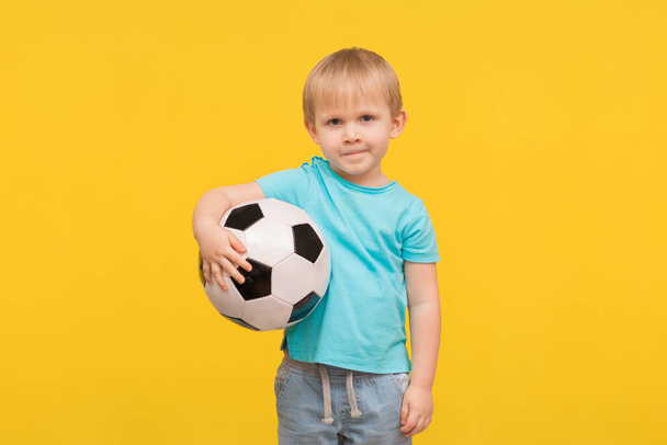A blond boy stands with a soccer ball and looks at the camera with a challenge and anticipation. A child on a spring bright yellow background with place for text, an idea for articles about sports. - Photo, Image