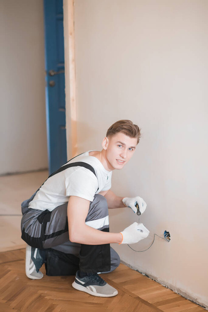 Young smiling electrical technician is connecting a mechanism of socket to wires in a white wall, using a screwdriver, repair works in a new apartment - Photo, image