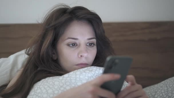 Handheld shot of woman scrolling a phone lying in bed - 映像、動画