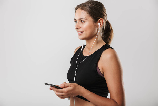 Image of beautiful woman in sportswear holding smartphone and listening to music with earphones isolated over white background - Fotoğraf, Görsel