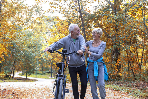 Ώριμο ζευγάρι γυναίκα και άνδρας με ποδήλατο περπατώντας στο πάρκο και να μιλήσουμε - Φωτογραφία, εικόνα