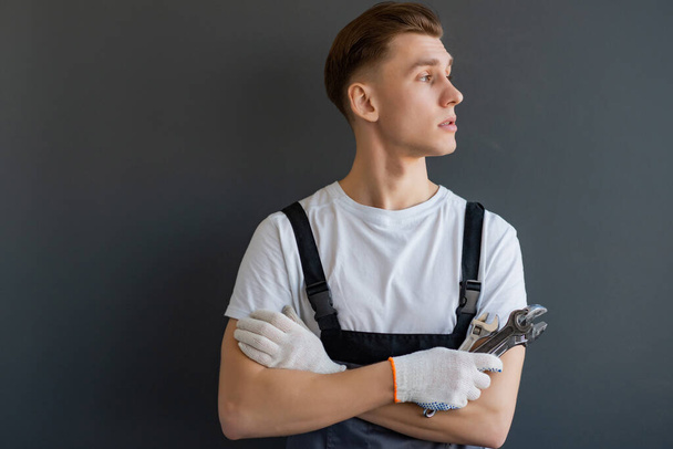 Young mechanic in overalls with crossed arms and wrench standing on gray background - Фото, зображення