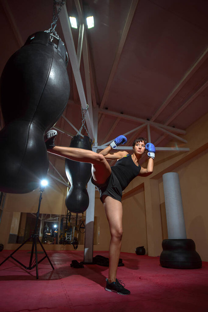Eine junge Kaukasierin trainiert aktiv im Fitnessstudio und macht Boxübungen in Boxhandschuhen vor einem Boxsack. Sport, Training, Motivation, aktives Lebensstilkonzept. - Foto, Bild