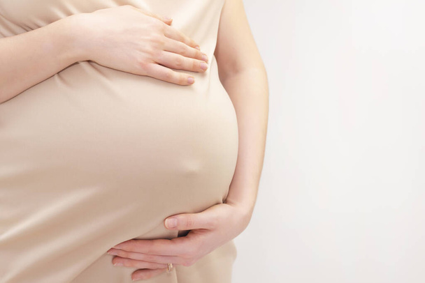 pregnant woman in a dress holds her hands on her stomach on a light background. Pregnancy, motherhood, preparation and expectation concept. Close-up indoors. Beautiful tender mood photo pregnancy. - Φωτογραφία, εικόνα