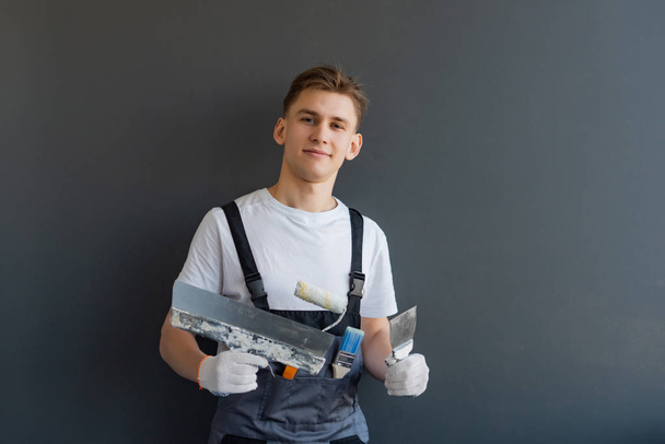 Mladý usměvavý muž v montérkách drží v ruce špachtle na šedém pozadí - Fotografie, Obrázek