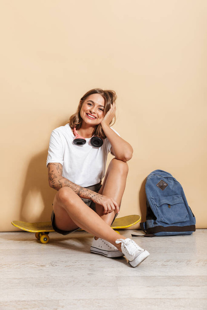 Portrait of a smiling schoolgirl sitting on a skateboard isolated over beige background, relaxing - Foto, immagini