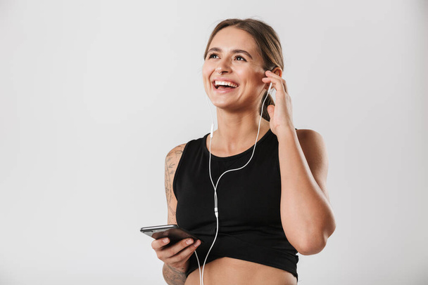 Image of caucasian woman in sportswear holding smartphone and listening to music with earphones isolated over white background - 写真・画像