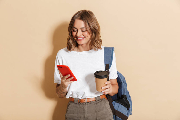 Image of caucasian student girl holding takeaway coffee cup and smartphone isolated over beige background - Фото, изображение