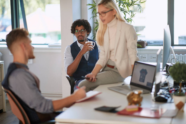 young female colleague sitting on desk, talking to one of the employees, while third one is watching, listening, analyzing information. business, casual, work, concept - Foto, afbeelding