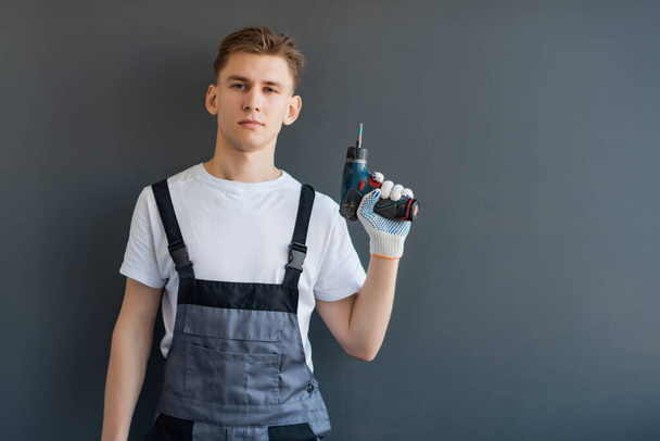 Portrét mladého usměvavého pracujícího muže v šedých kombinézách a bílém tričku. Pracovník drží elektrický vrták v ruce na šedém pozadí.místo pro text, kopírovat prostor - Fotografie, Obrázek