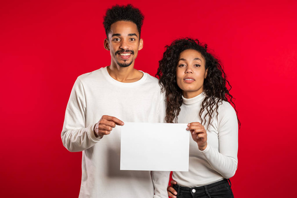 Portrait of young handsome african american man and woman couple holding white empty paper blank on red studio background. Copy space. Match with sign - Fotografie, Obrázek