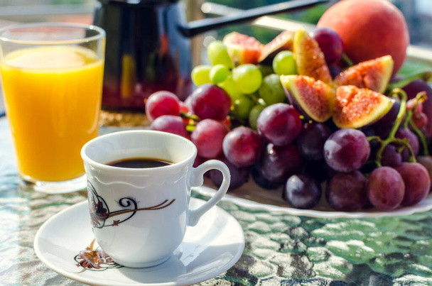 Frühstück im Freien auf dem Tisch mit einer Tasse Kaffee und türkischer Kaffeekanne, einem Glas frischem Orangensaft, einem Teller mit weißen und weinroten Trauben, Feigen, Pfirsich. Eine Tasse Kaffee im Vordergrund. - Foto, Bild