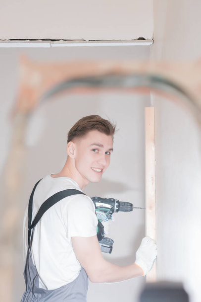 Smiling young working man in overalls drills screws with an electric screwdriver in an apartment, home improvement concept - Фото, изображение
